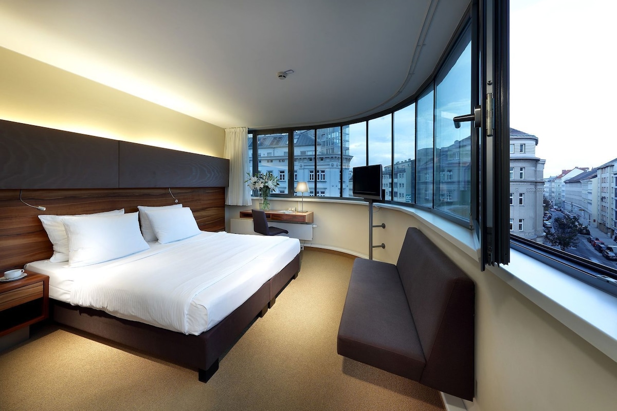 A modern hotel room with a large bed, a desk, a chair, and a large window offering a city view.