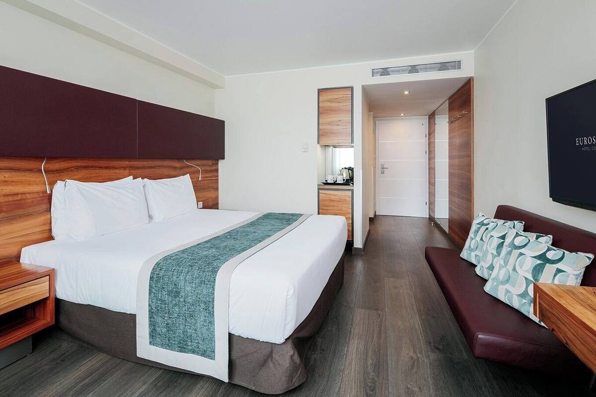A modern hotel room with a large bed, a wooden headboard, a bedside table, a bench with patterned cushions, and a flat-screen TV mounted on the wall.