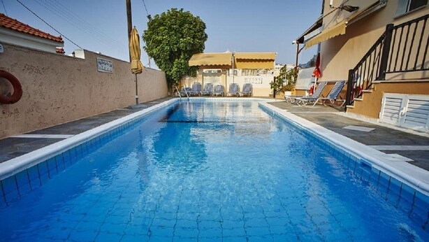 A swimming pool with tiled flooring, sun loungers, and a building with a tiled roof in the background. - Angela Apartments (Samos)
