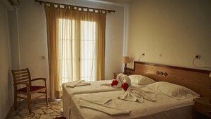 A hotel room with two beds, a wooden chair, a table, and a window with curtains. - Angela Apartments (Samos)