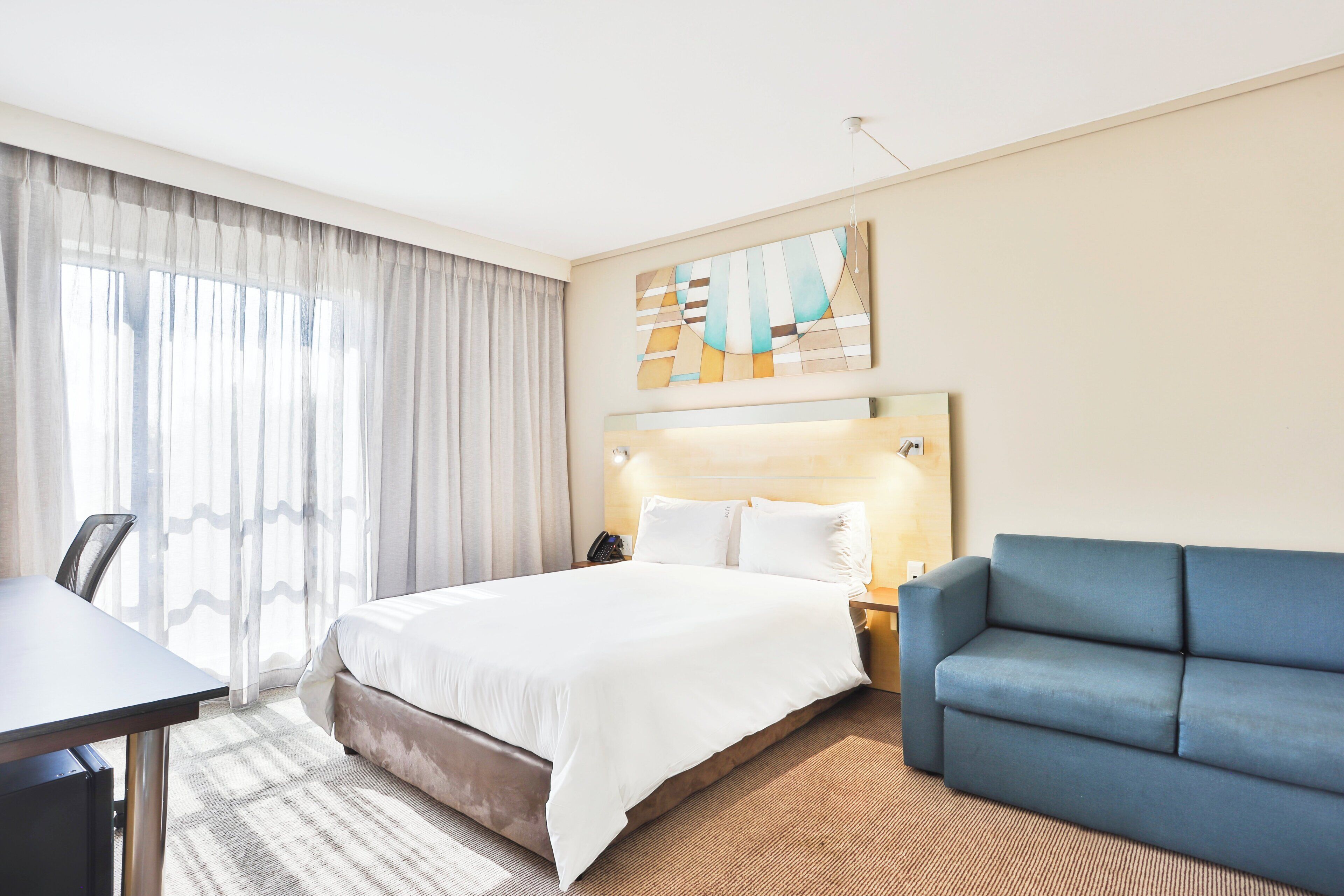 Una habitación de hotel con cama, escritorio, sofá azul y un cuadro en la pared.