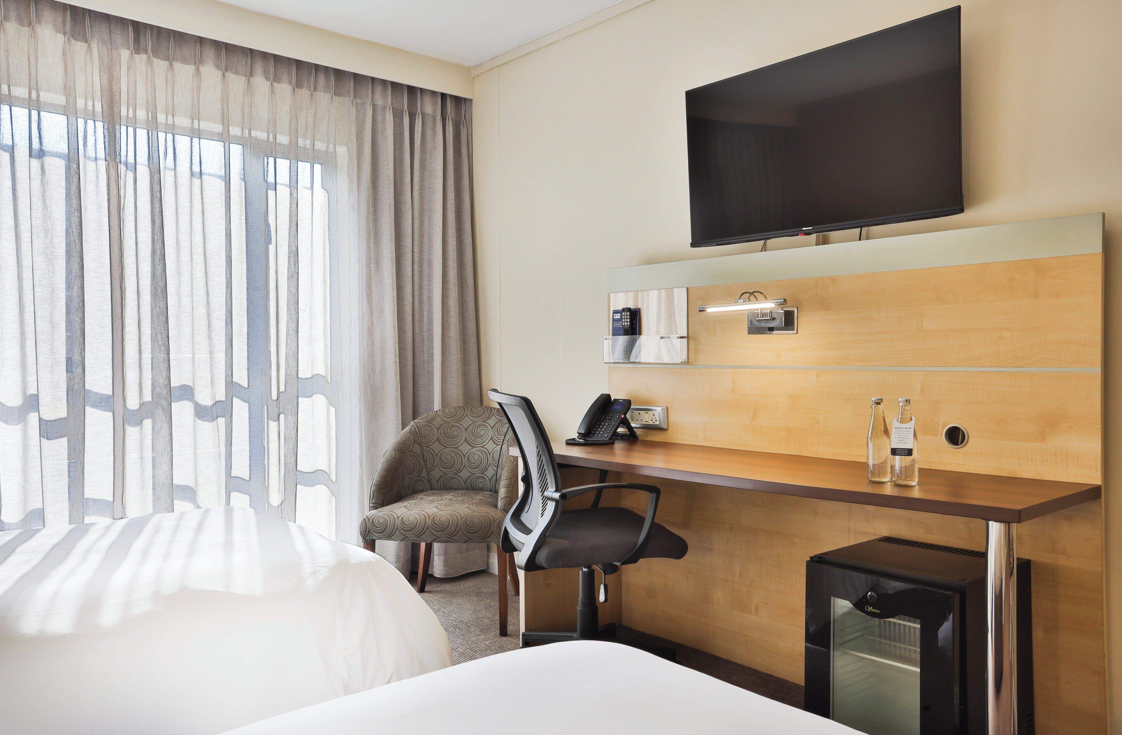 Habitación de hotel con cama, escritorio con teléfono y cafetera, una silla, televisor montado en la pared y ventana con cortinas.