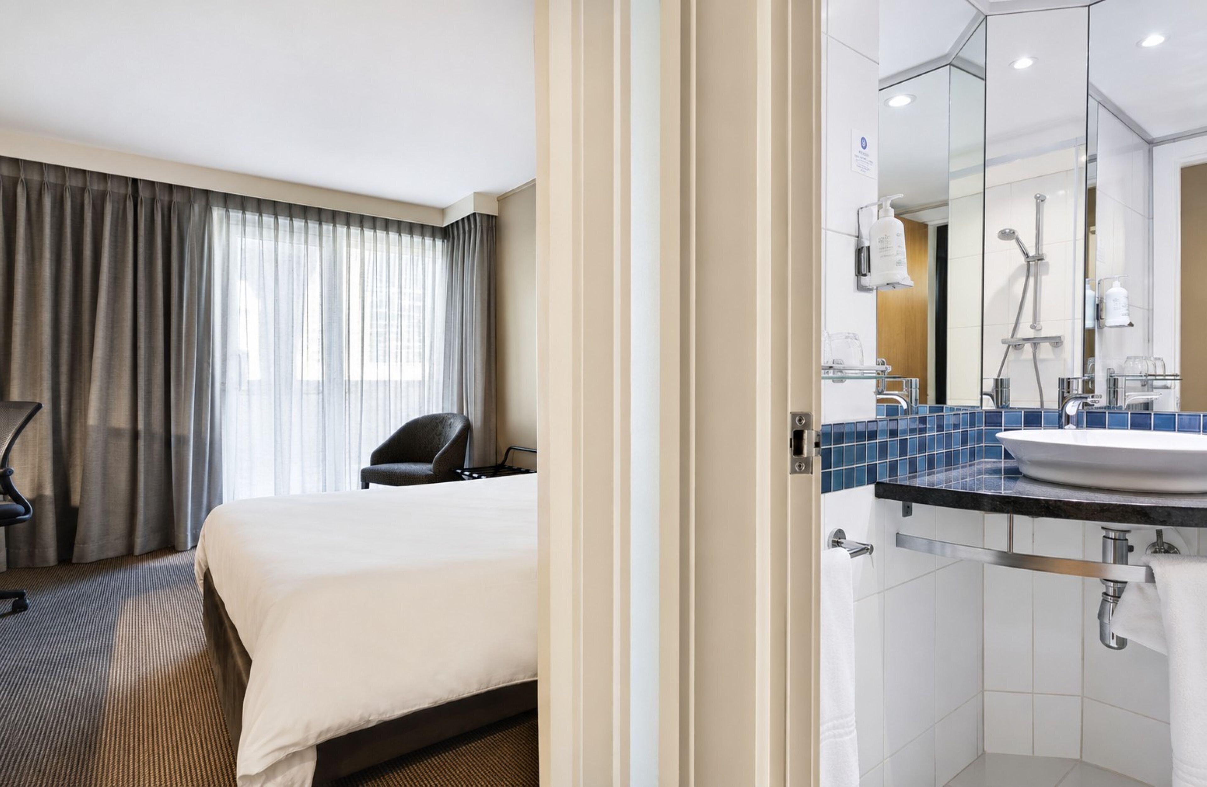 A hotel room with a bed, a chair, a bathroom with a sink and mirror, and a shower.