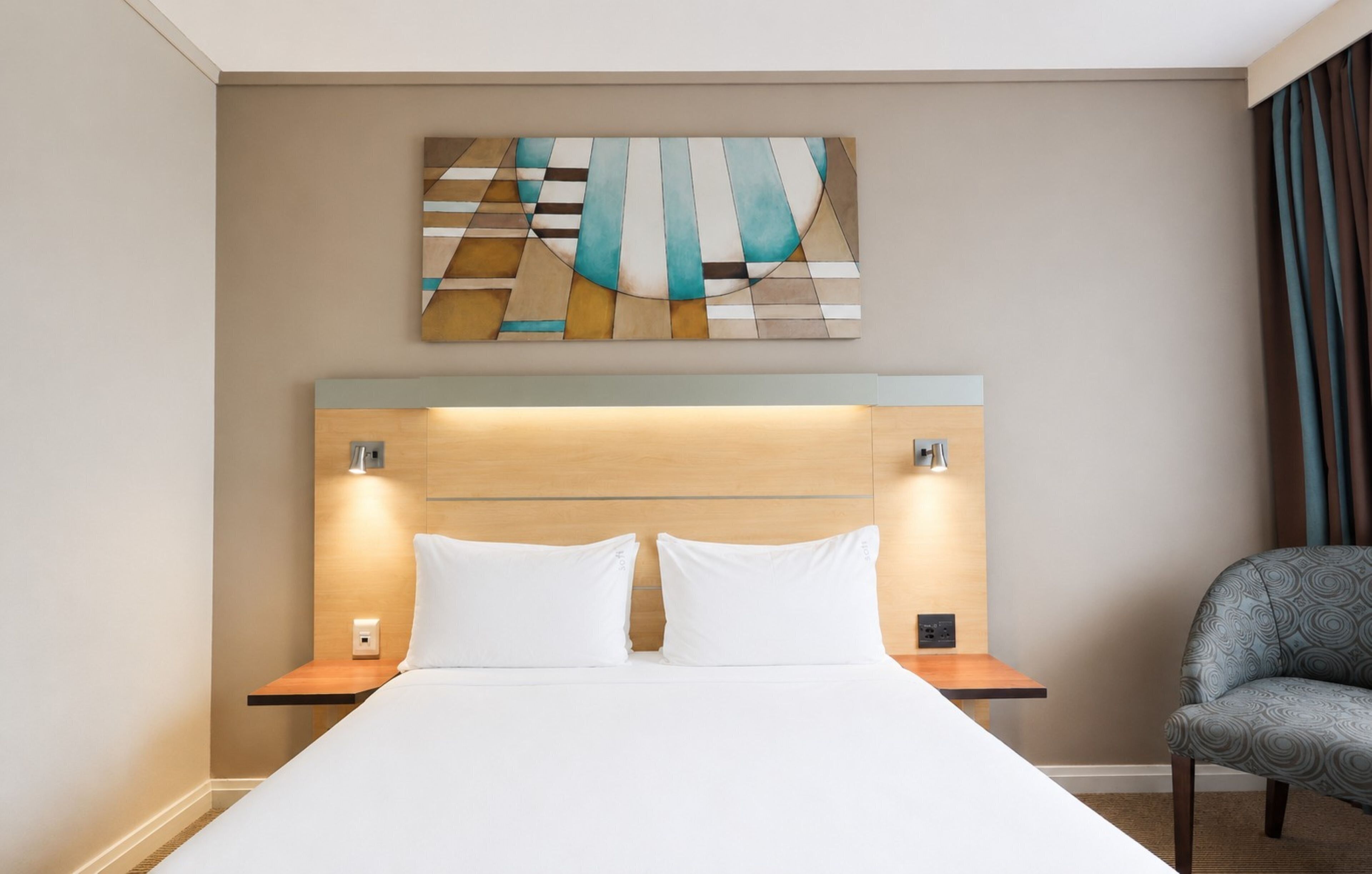 Habitación de hotel con cama, dos mesitas de noche, una silla y un cuadro abstracto colgado en la pared.