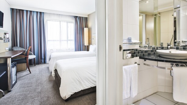 A hotel room with two beds, a desk, a chair, a large window with curtains, and a bathroom with a sink and mirror. - Holiday Inn Express Sandton Woodmead by IHG (Sandton)