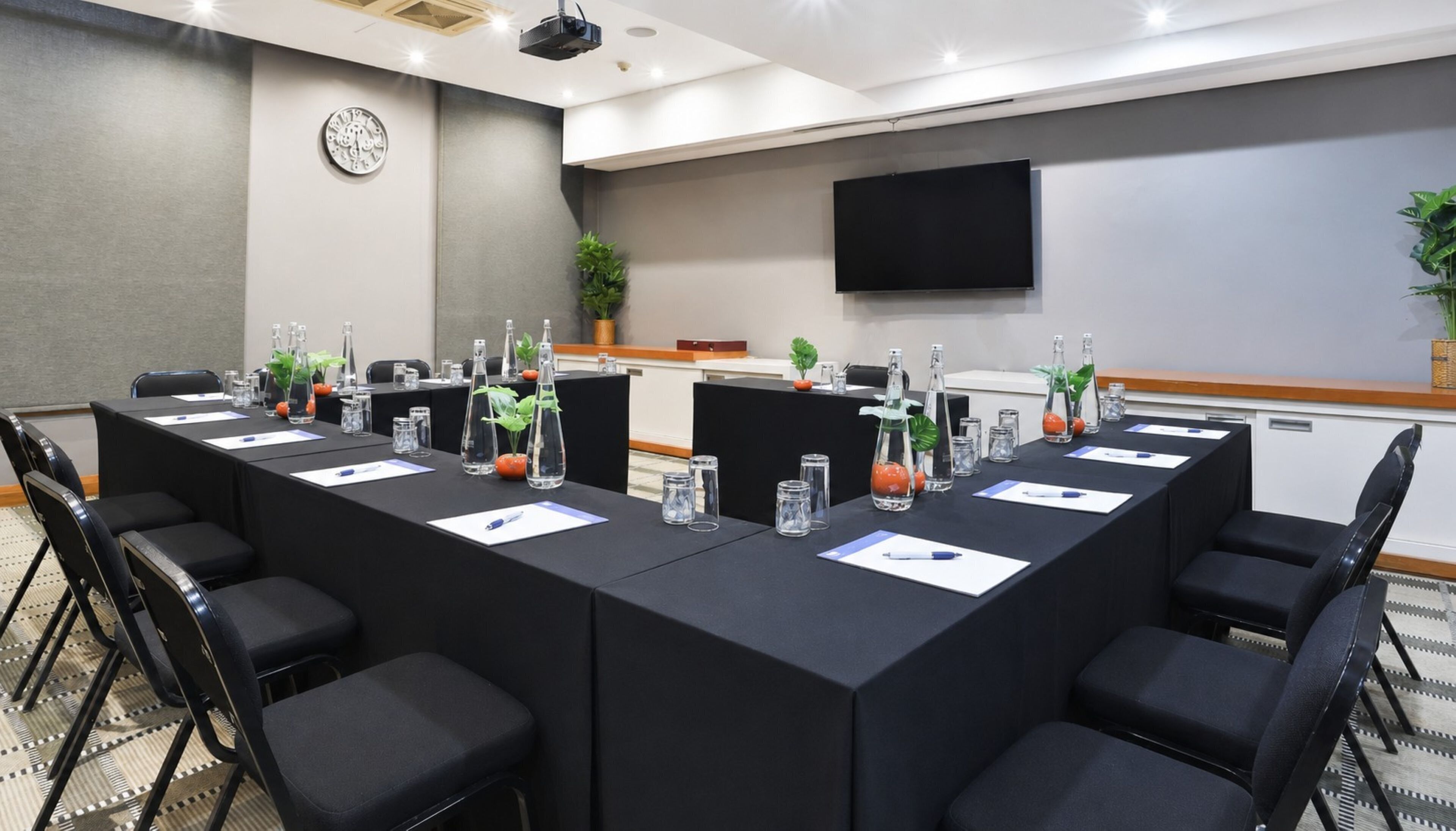 Un salón de conferencias con una mesa larga, sillas, botellas de agua y un reloj en la pared.