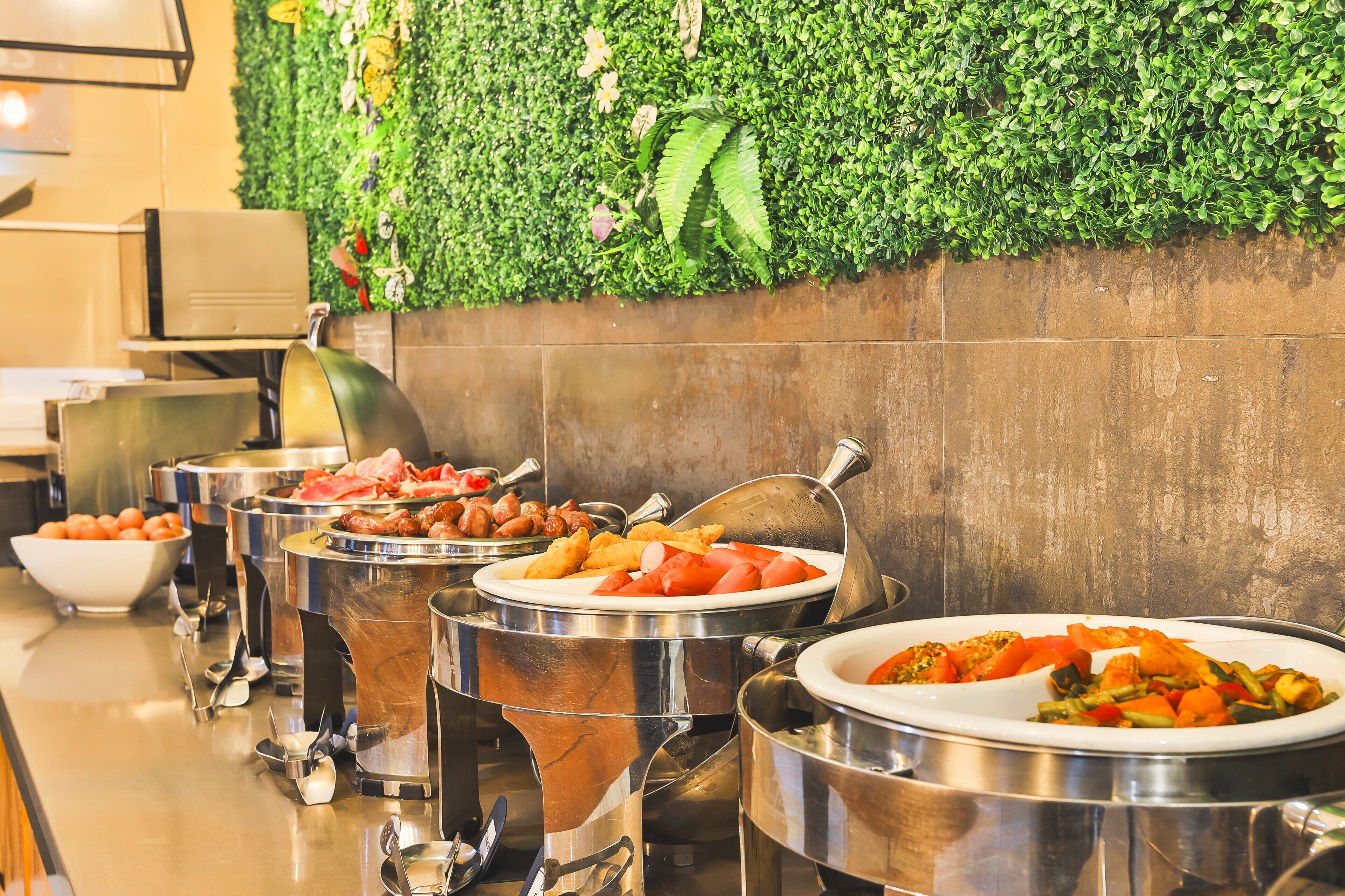 Una mesa de buffet con diversos alimentos en recipientes metálicos, una pared verde con follaje y un suelo de baldosas.