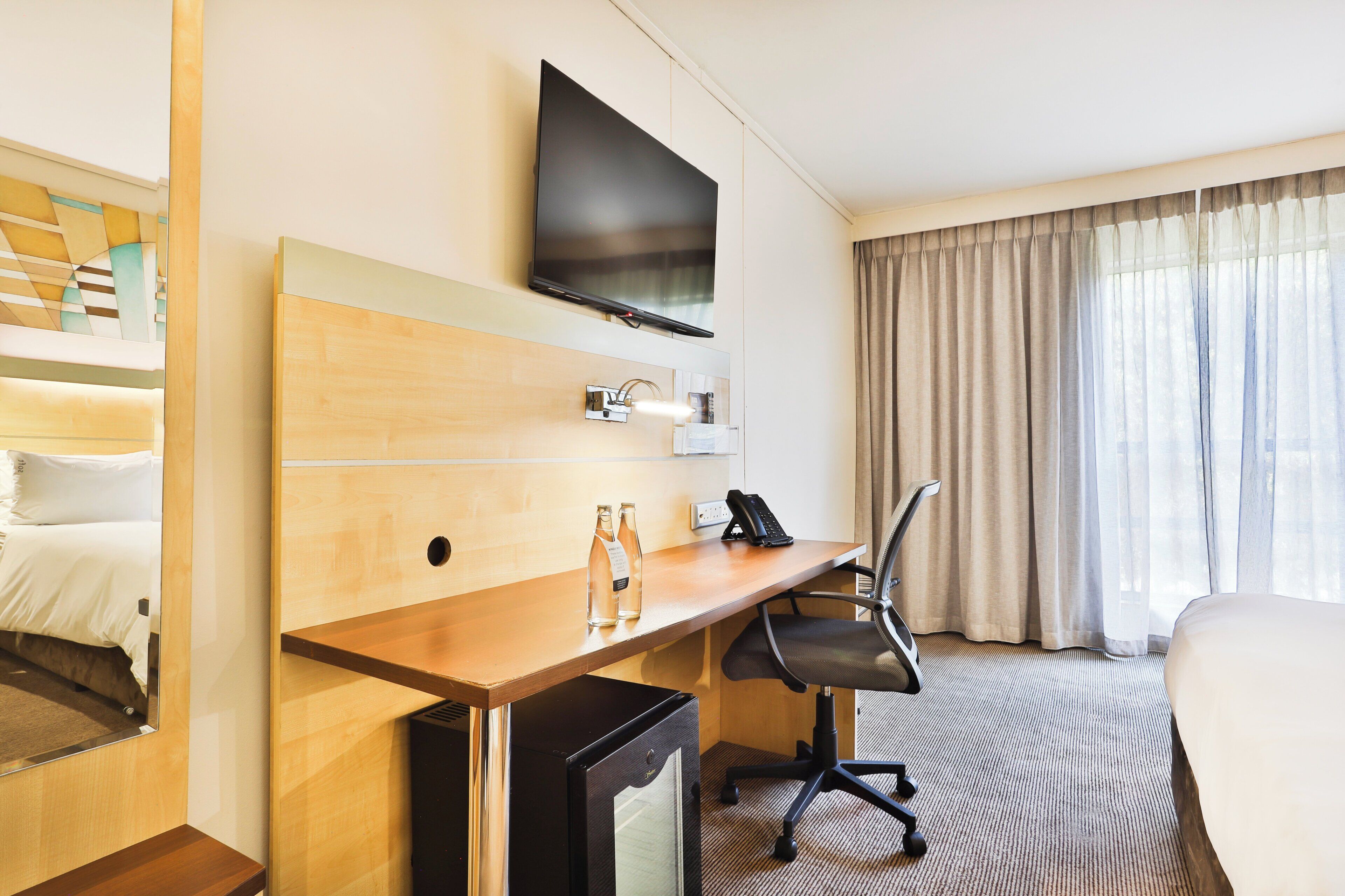 Habitación de hotel con cama, escritorio con silla, televisor montado en la pared y ventana con cortinas.