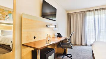 Habitación de hotel con cama, escritorio con silla, televisor montado en la pared y ventana con cortinas.