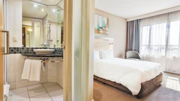 Un baño de hotel con lavamanos, espejo y ducha. La habitación tiene una cama con ropa de cama blanca y una silla.