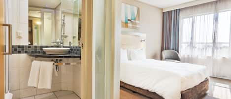 A hotel bathroom with a sink, mirror, and shower. The room has a bed with white bedding and a chair.