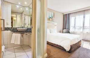 A hotel bathroom with a sink, mirror, and shower. The room has a bed with white bedding and a chair. - Holiday Inn Express Sandton Woodmead by IHG (Sandton)