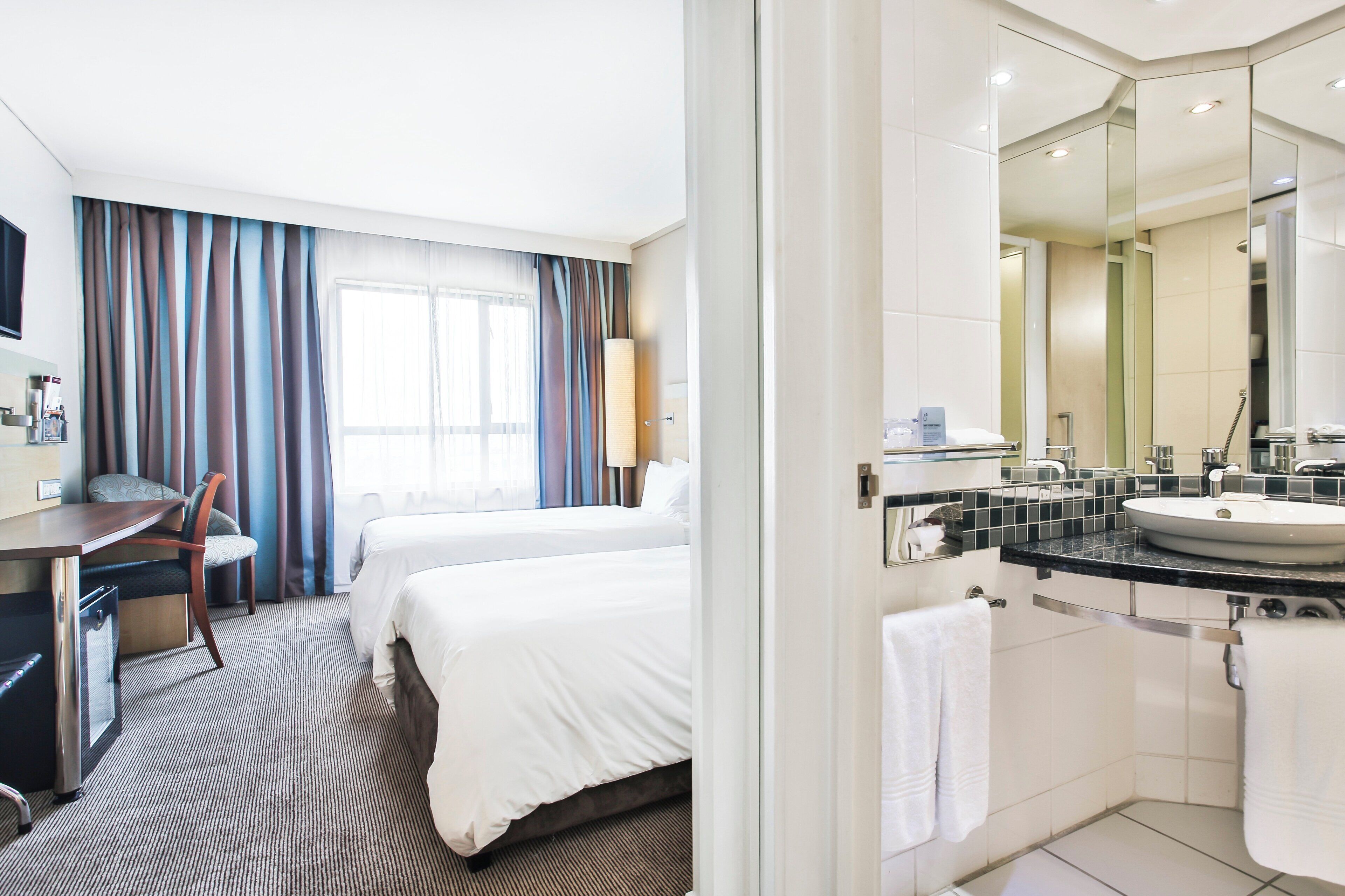 A hotel room with a bed, a desk, a chair, a bathroom with a sink and mirror, and a window with curtains.