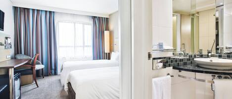 A hotel room with a bed, a desk, a chair, a bathroom with a sink and mirror, and a window with curtains.