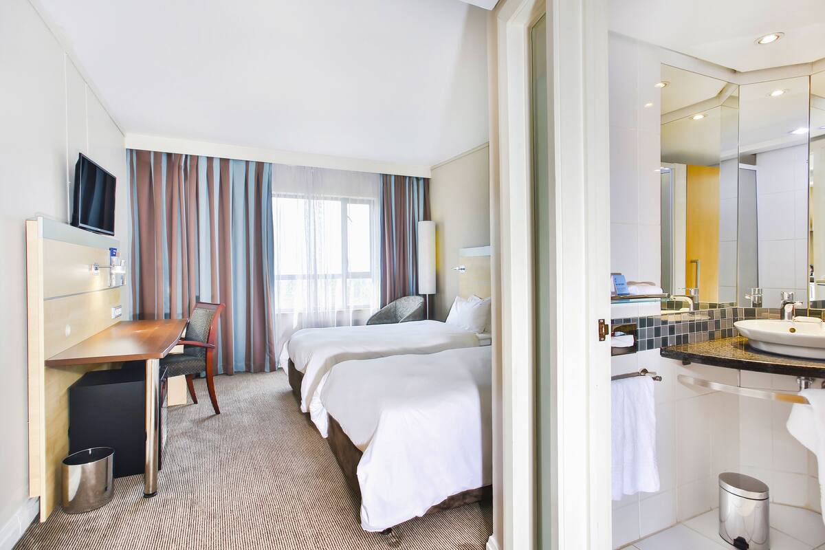 A hotel room with two beds, a desk, a chair, a TV, a bathroom with a sink and mirror, and a trash bin.