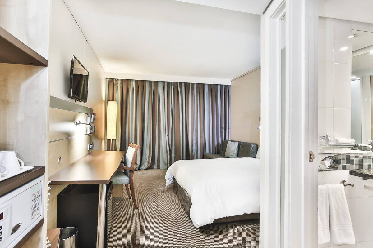 A hotel room with a bed, a desk with a chair, a television, and a bathroom with a mirror and towel rack.