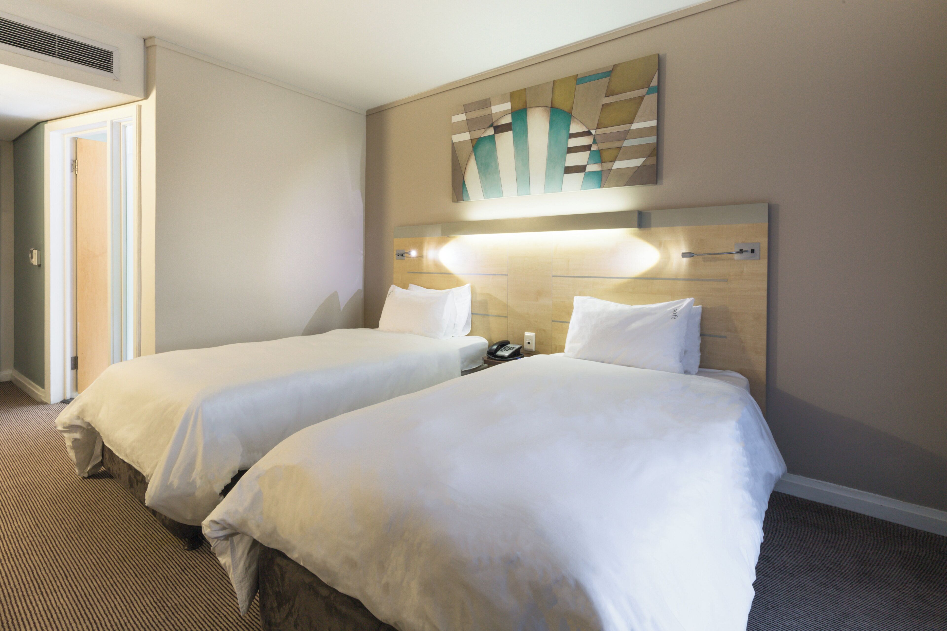 Habitación de hotel con dos camas, cabecera con iluminación integrada, teléfono y una obra de arte colgada en la pared.