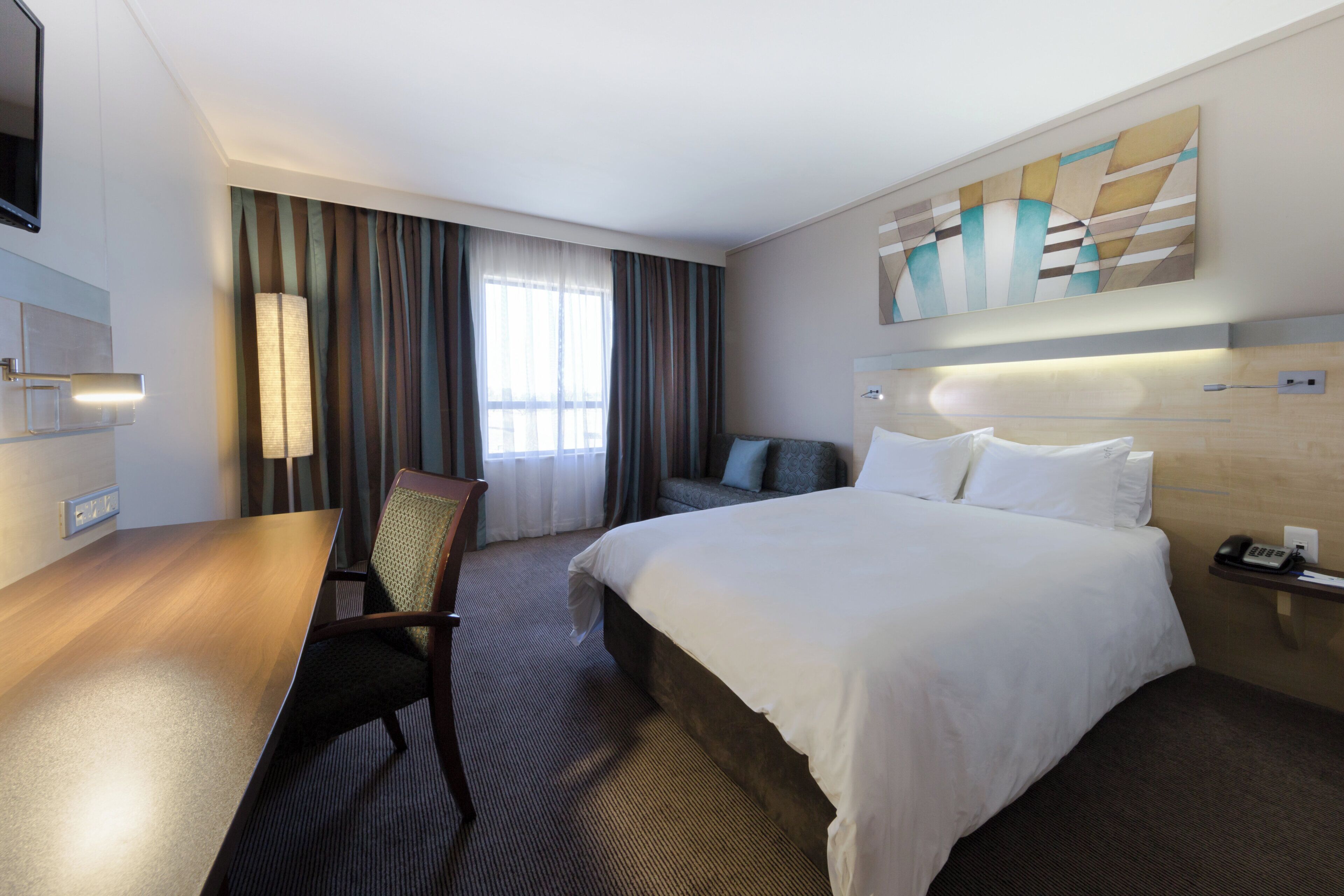 A hotel room with a bed, a desk, a chair, a sofa, a painting on the wall, and a window with curtains.