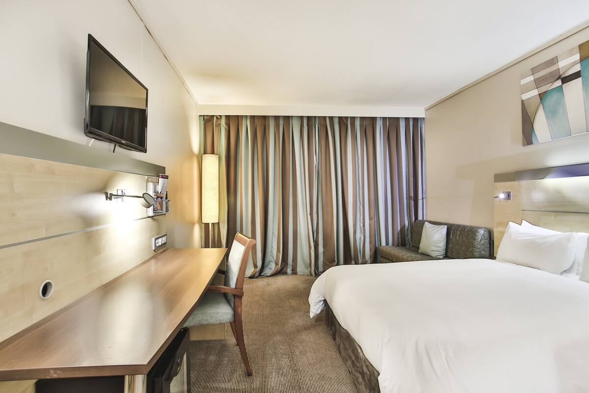 A hotel room with a bed, a desk, a chair, a TV mounted on the wall, and a curtain backdrop.