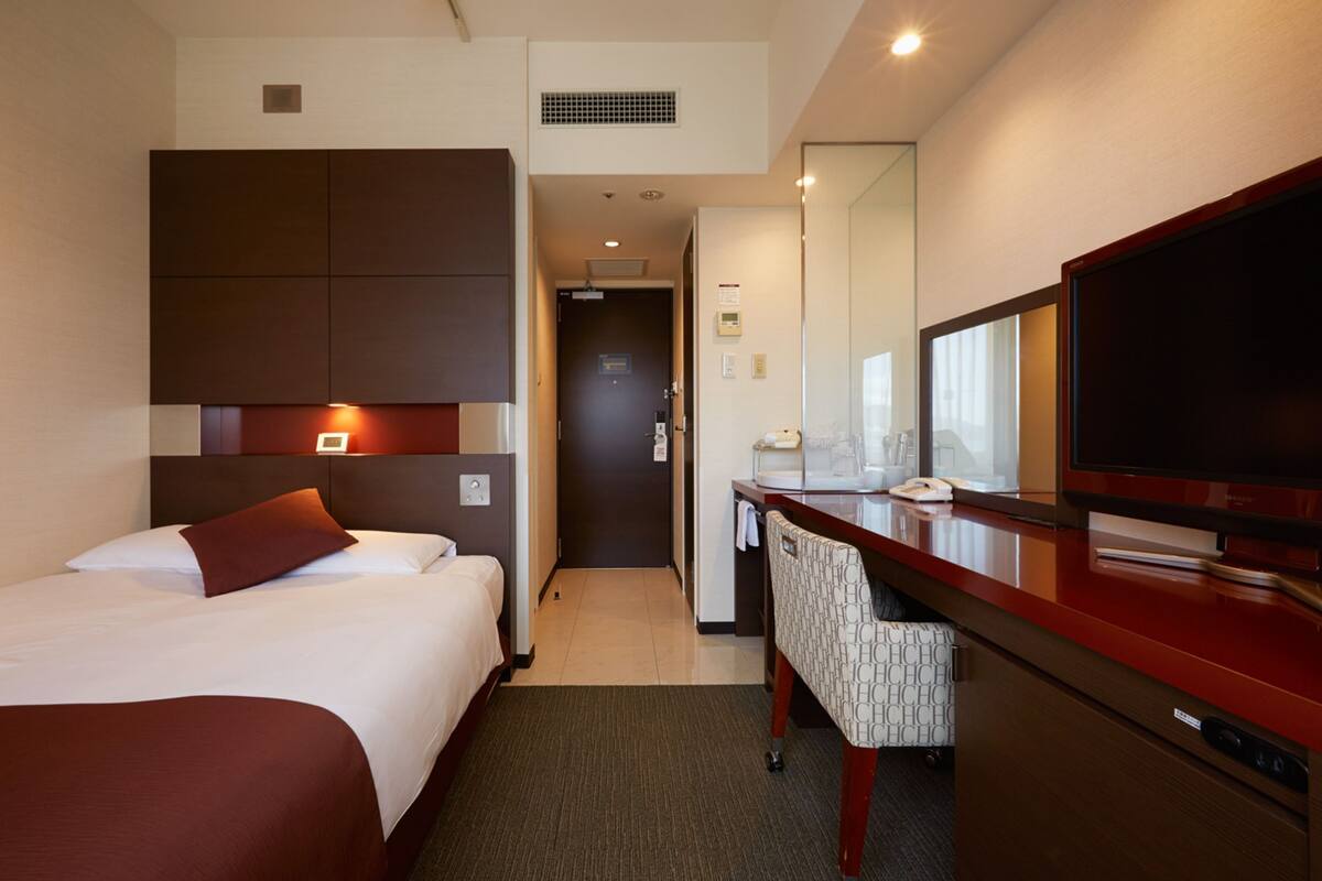 Una habitación de hotel con cama, escritorio, silla, televisión y espejo.