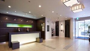 A modern reception area with a reception desk, two staff members, a green illuminated panel, and a large window with a view of the outdoors.