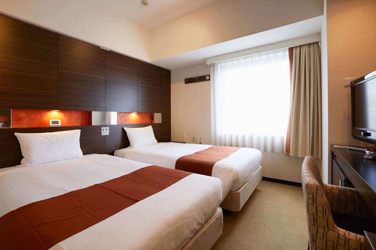 Habitación de hotel con dos camas, televisión, escritorio y ventana con cortinas.
