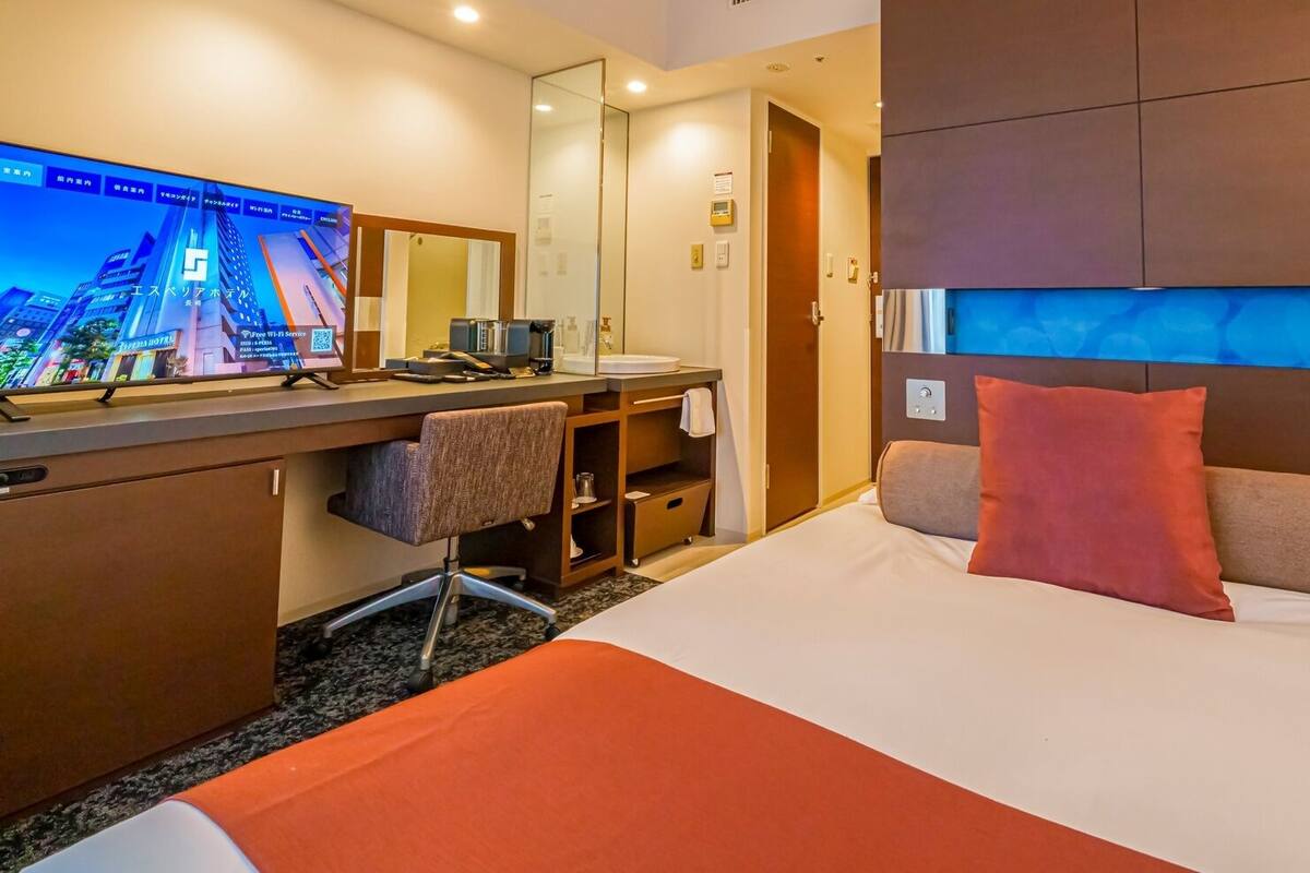 Habitación de hotel con cama, escritorio, silla y un televisor grande que muestra un paisaje urbano.