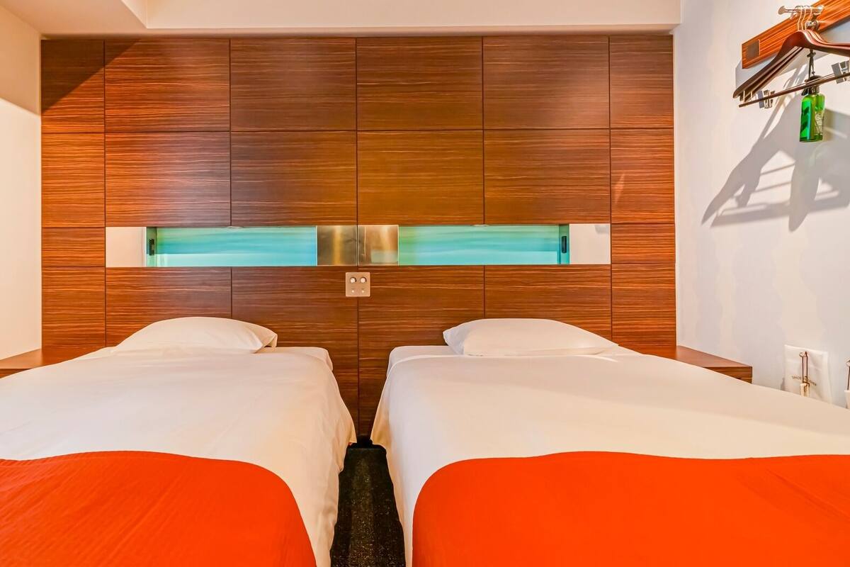 Habitación de hotel con dos camas, cabecera de madera, estante empotrado y perchero.