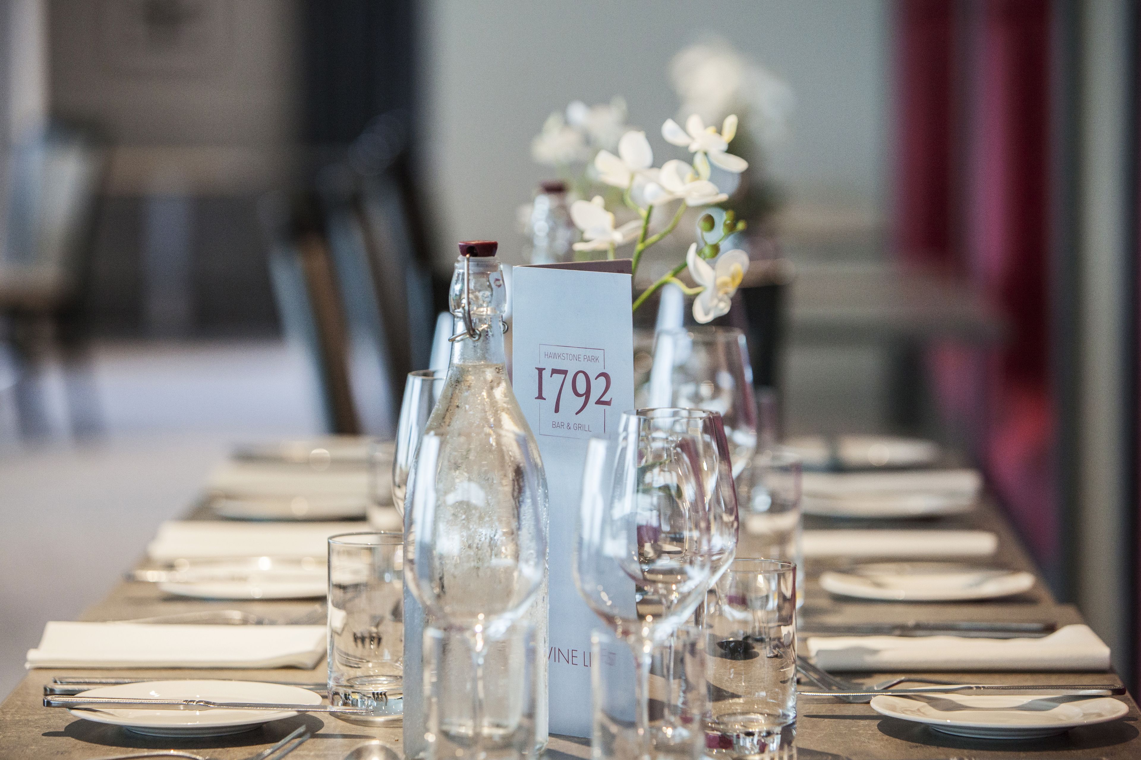 Een gedekte restauranttafel met borden, bestek, glazen en een waterfles. Een bord met het nummer 1792 is zichtbaar.