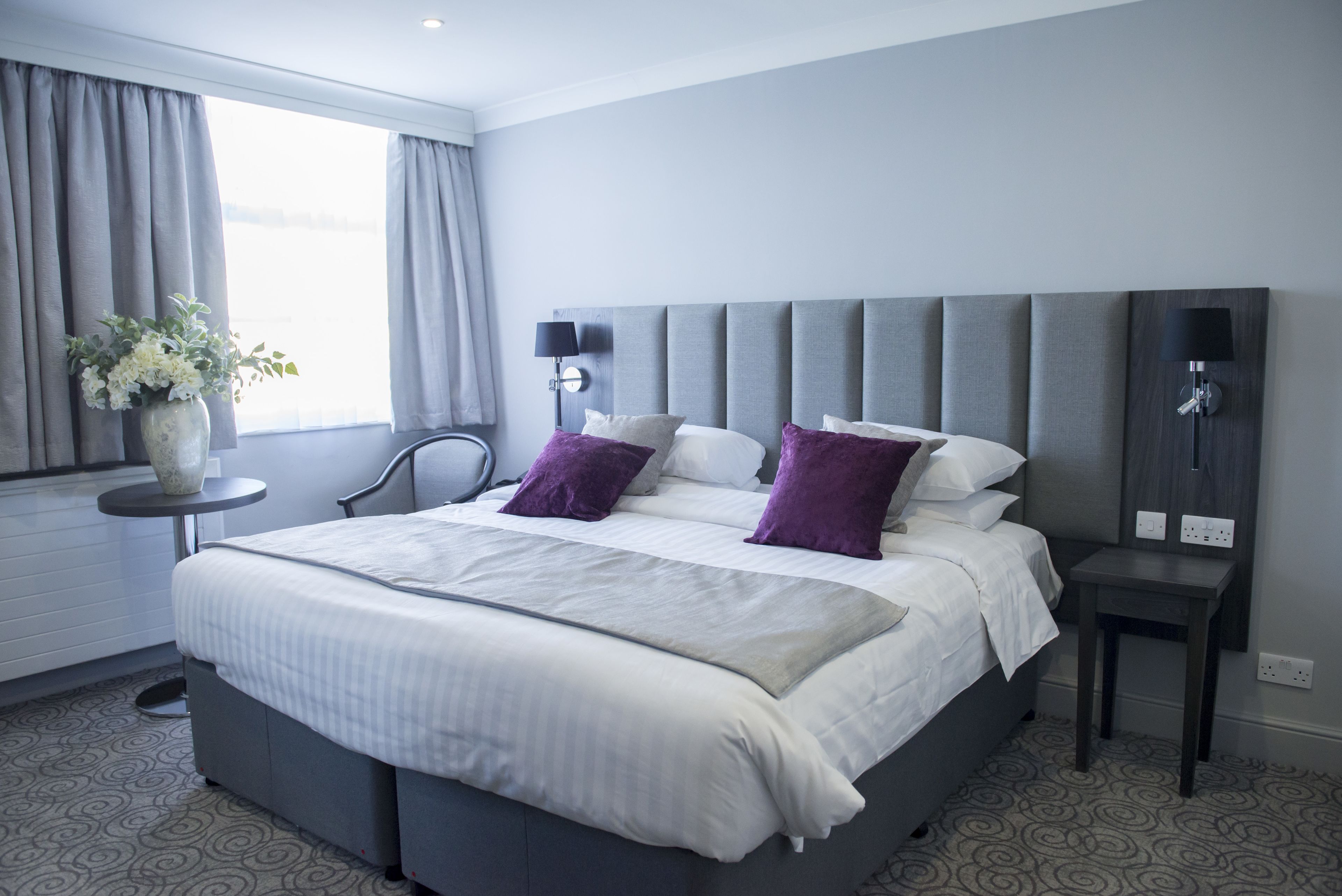 A modern hotel room with a large bed, a side table, a lamp, and a window with curtains.