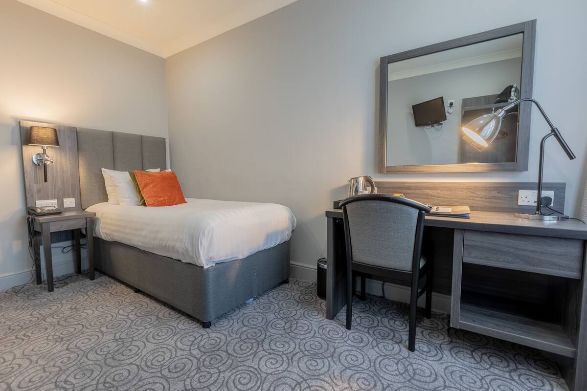A hotel room with a bed, a desk, a chair, a desk lamp, a TV, and a trash bin.