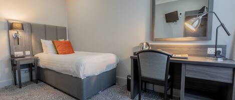 A hotel room with a bed, a desk, a chair, a desk lamp, a TV, and a trash bin.