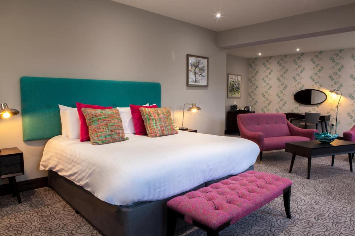 A modern hotel room with a large bed, a pink armchair, a small table, and a patterned carpet.
