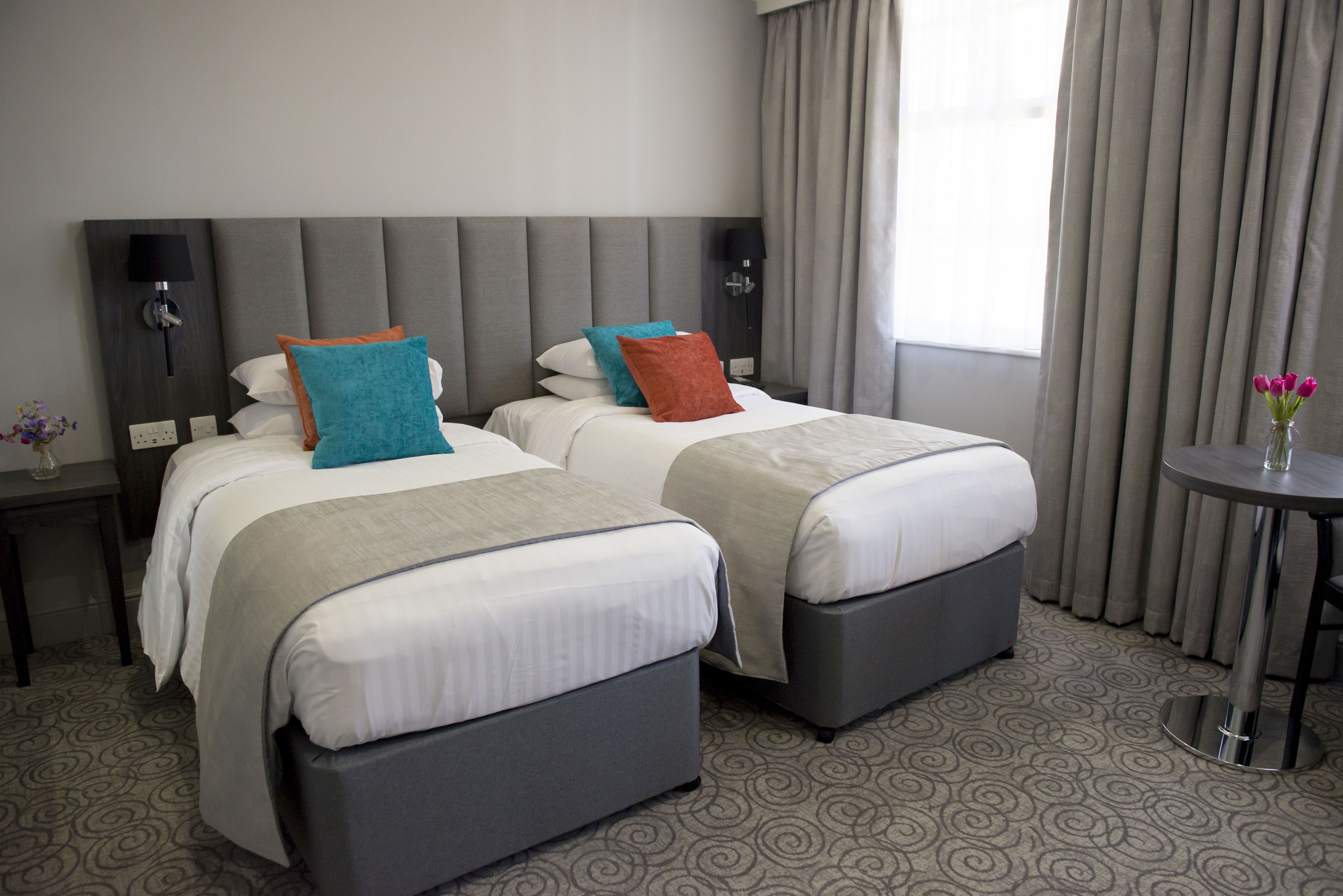 A hotel room with two beds, a small table, and a nightstand with a lamp.
