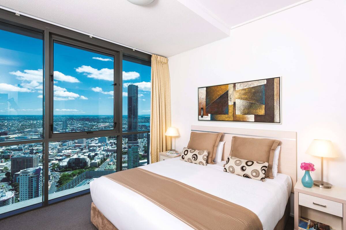 A hotel room with a large bed, a city view, a painting on the wall, and a nightstand with a lamp.