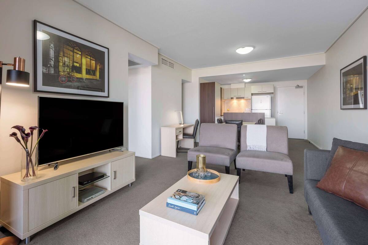 A modern living room with a flat-screen TV, a coffee table with a book, a grey sofa, and a grey armchair.