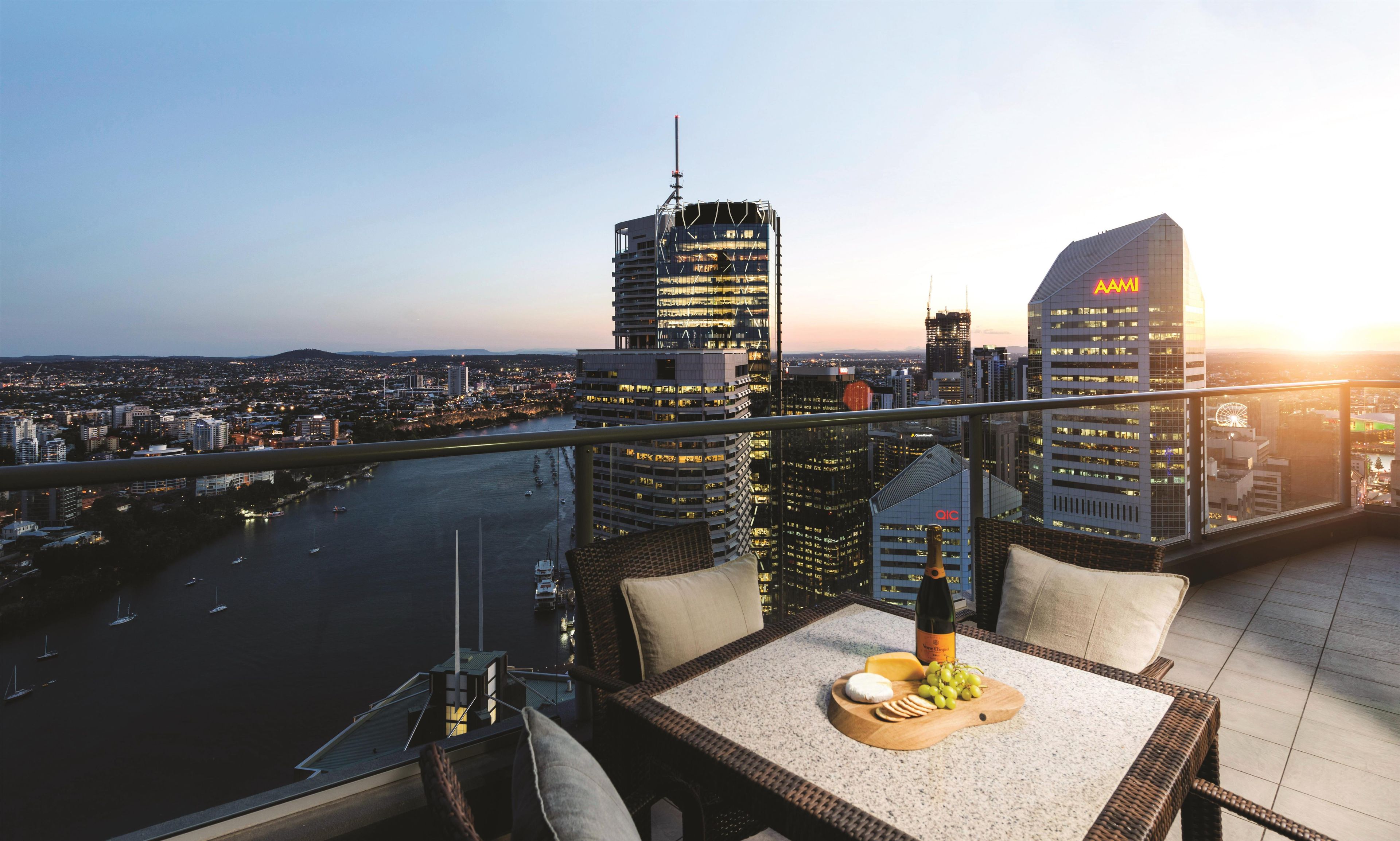 Un balcón con una mesa dispuesta para una comida, con vistas a un paisaje urbano que incluye edificios modernos y un río.