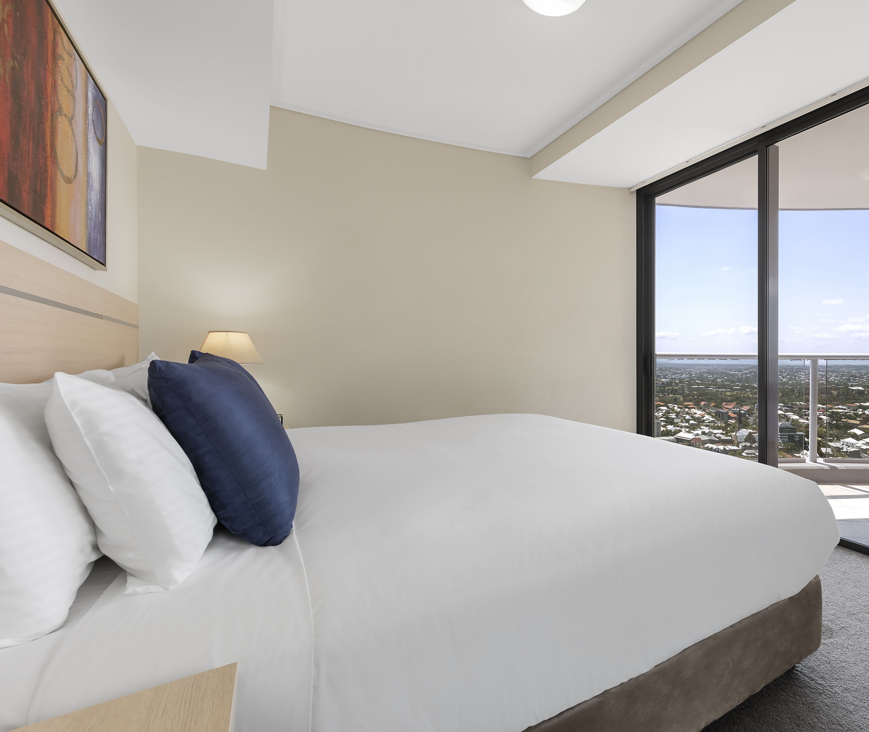 A hotel room with a large bed, a nightstand with a lamp, and a view of the city through large windows.