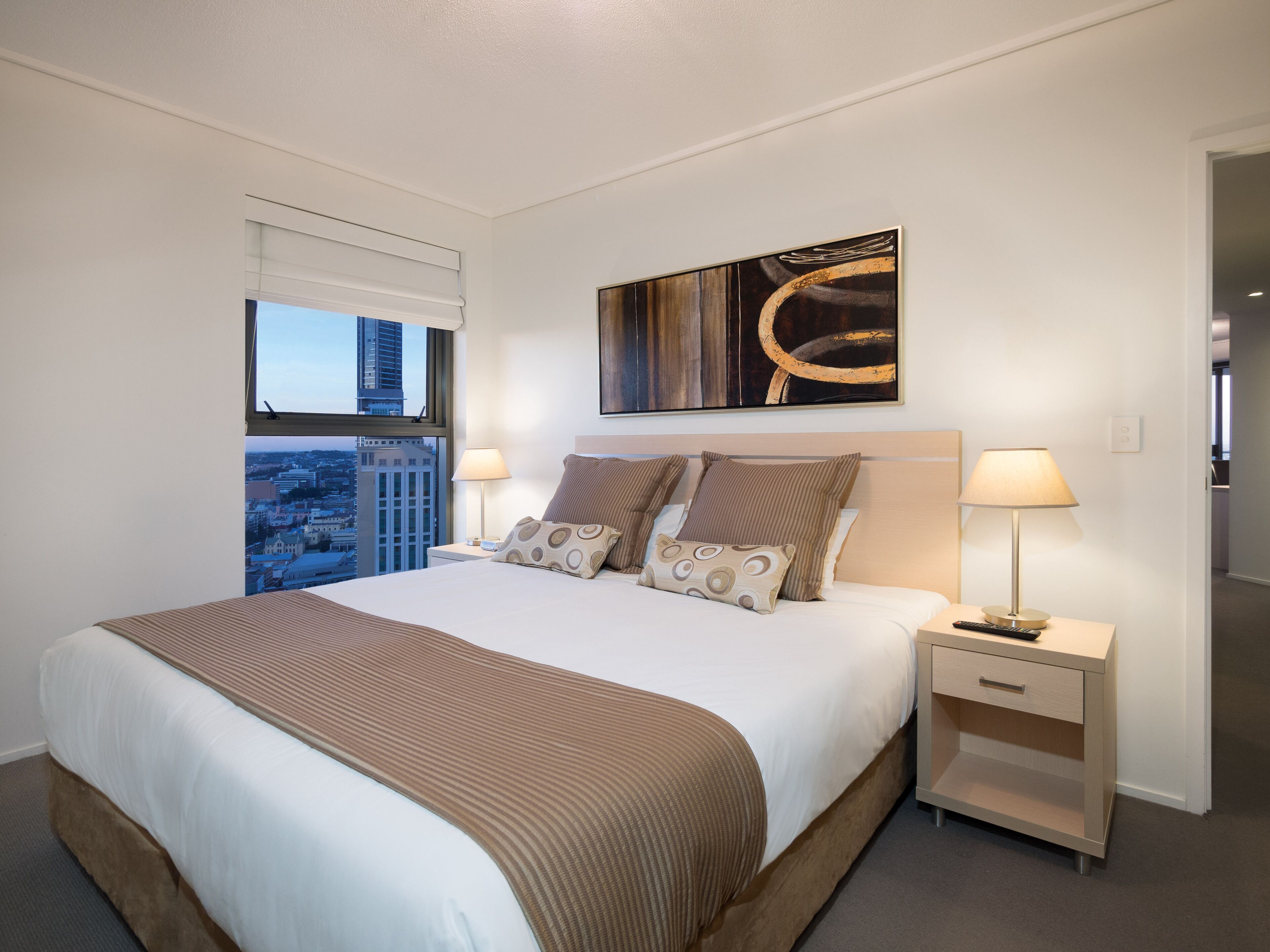 Quarto de hotel com uma cama grande, duas mesinhas de cabeceira, uma janela com vista para a cidade e um quadro na parede.
