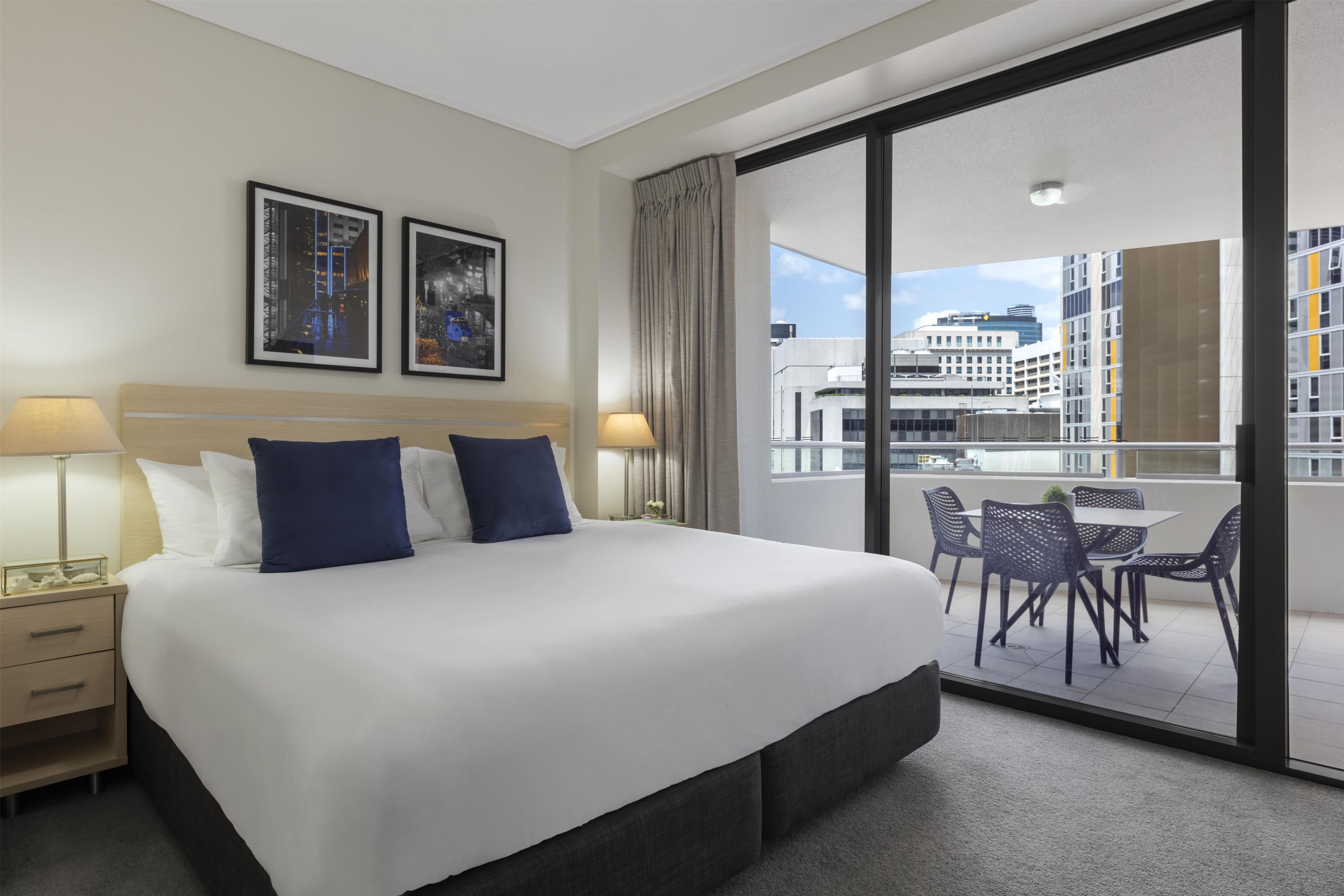 A hotel room with a large bed, two bedside lamps, a nightstand, a window with a view of the city, and a balcony with outdoor seating.