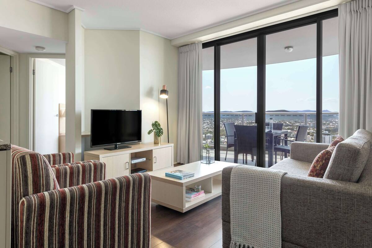 A modern living room with a flat-screen TV, striped sofas, a coffee table, and a large window offering a city view.