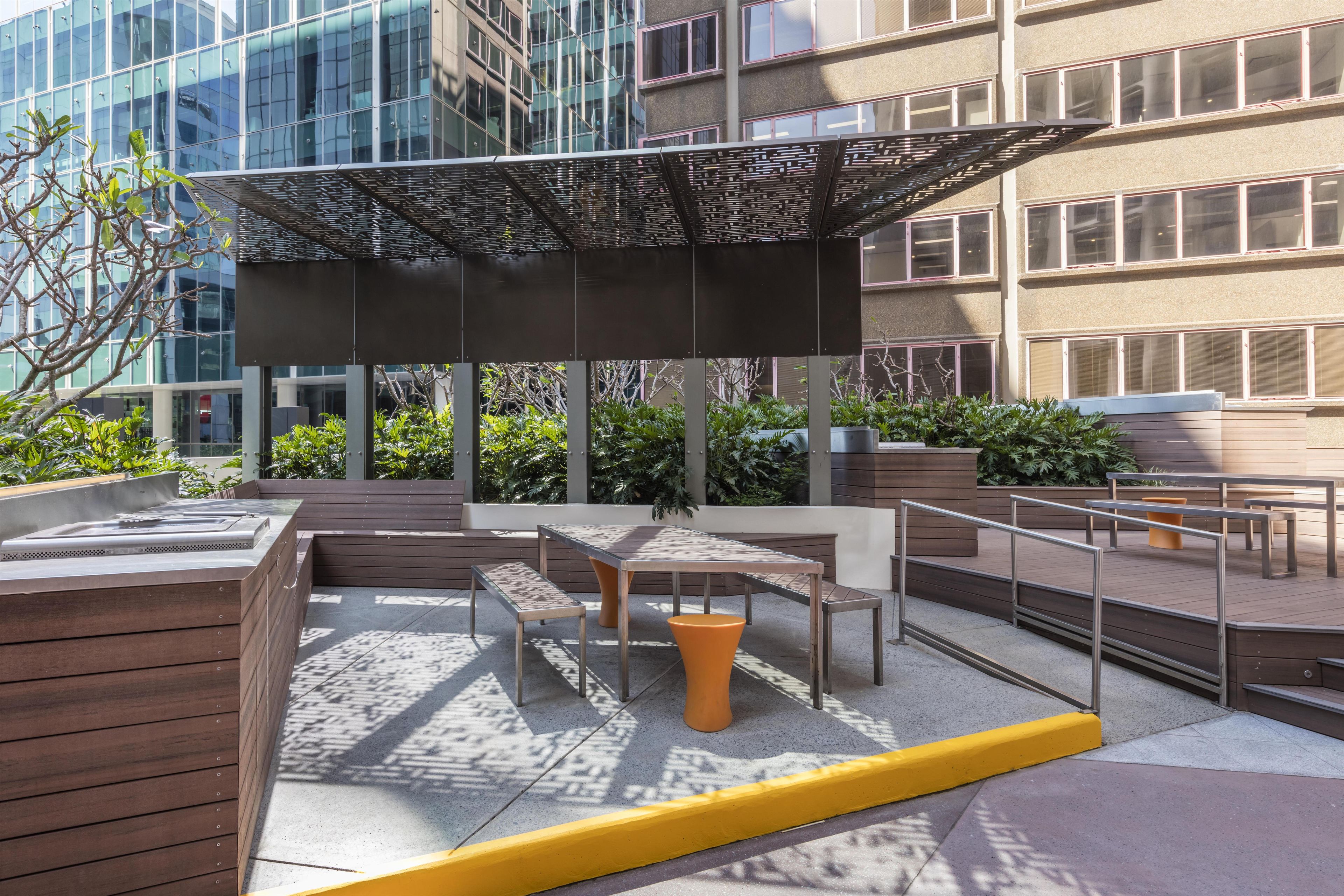 An outdoor seating area with tables and benches, surrounded by greenery and a modern building in the background.