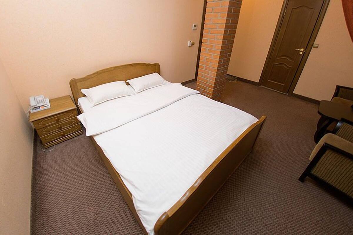 A neatly made bed in a hotel room with a wooden headboard, a nightstand with a lamp, and a small table with chairs.