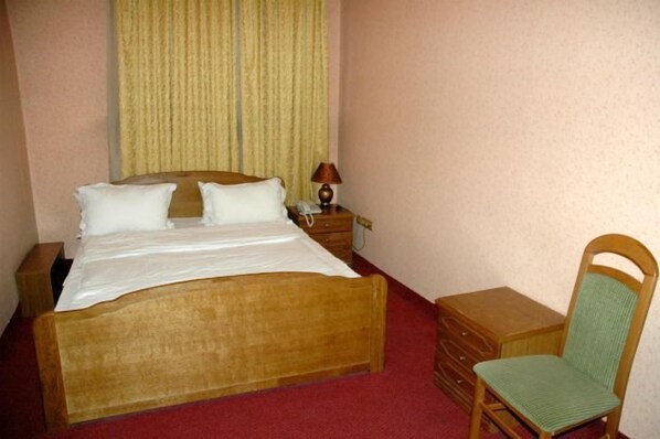 A single bed with a wooden headboard, a bedside table with a lamp, a chair, and a small dresser. - Almaty Sapar (Almaty)