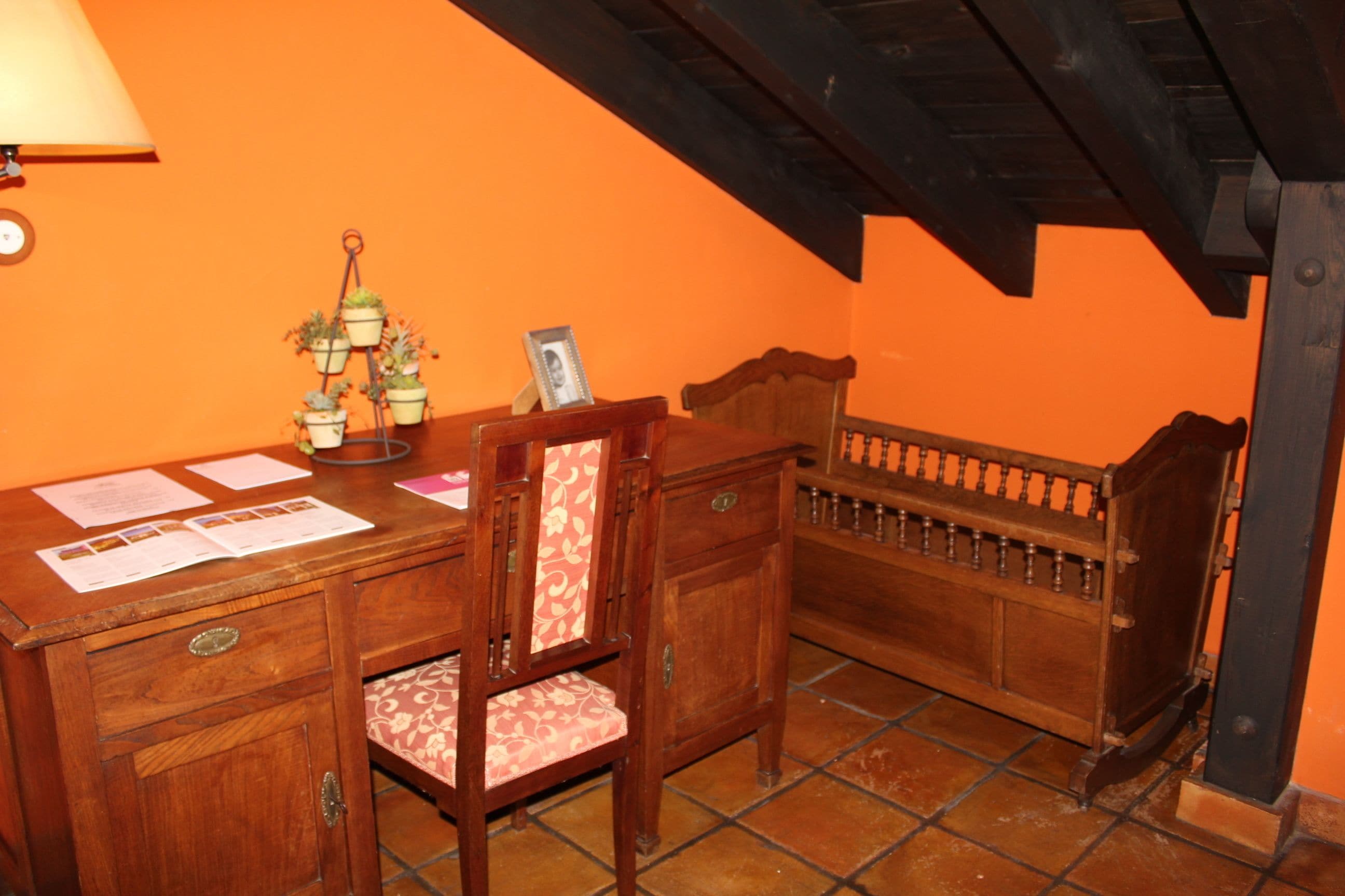 A wooden desk with papers, a lamp, and a chair in a room with orange walls and a tiled floor.