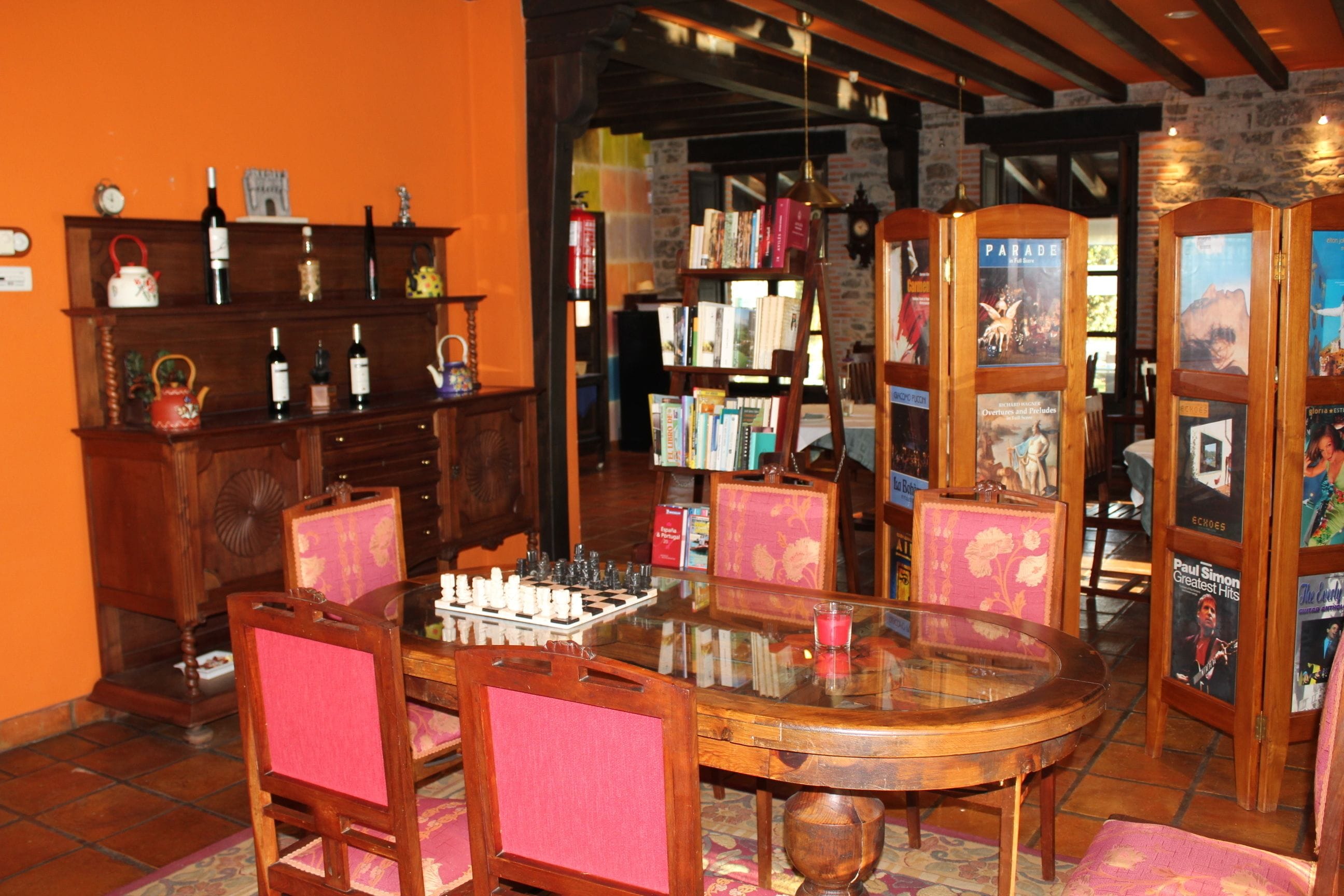 A cozy room with a wooden table, chairs, and a chessboard. There are bookshelves with books and decorative items.