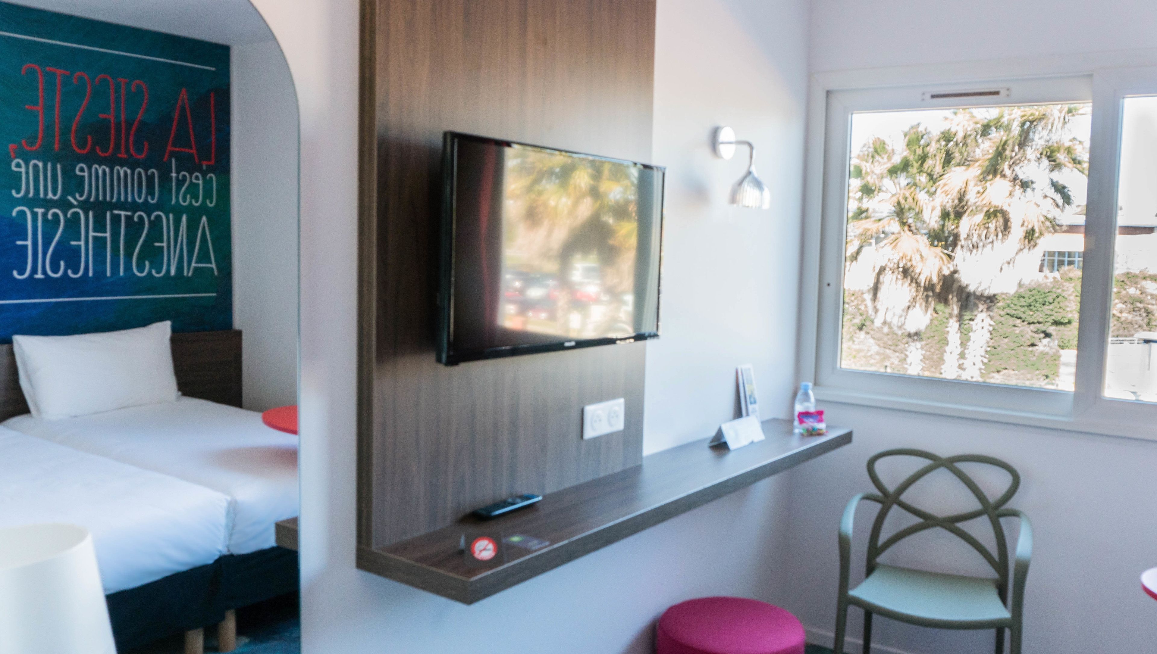 Habitación de hotel moderna con cama, televisor montado, una mesita con un control remoto, una silla y una ventana con vistas a palmeras.