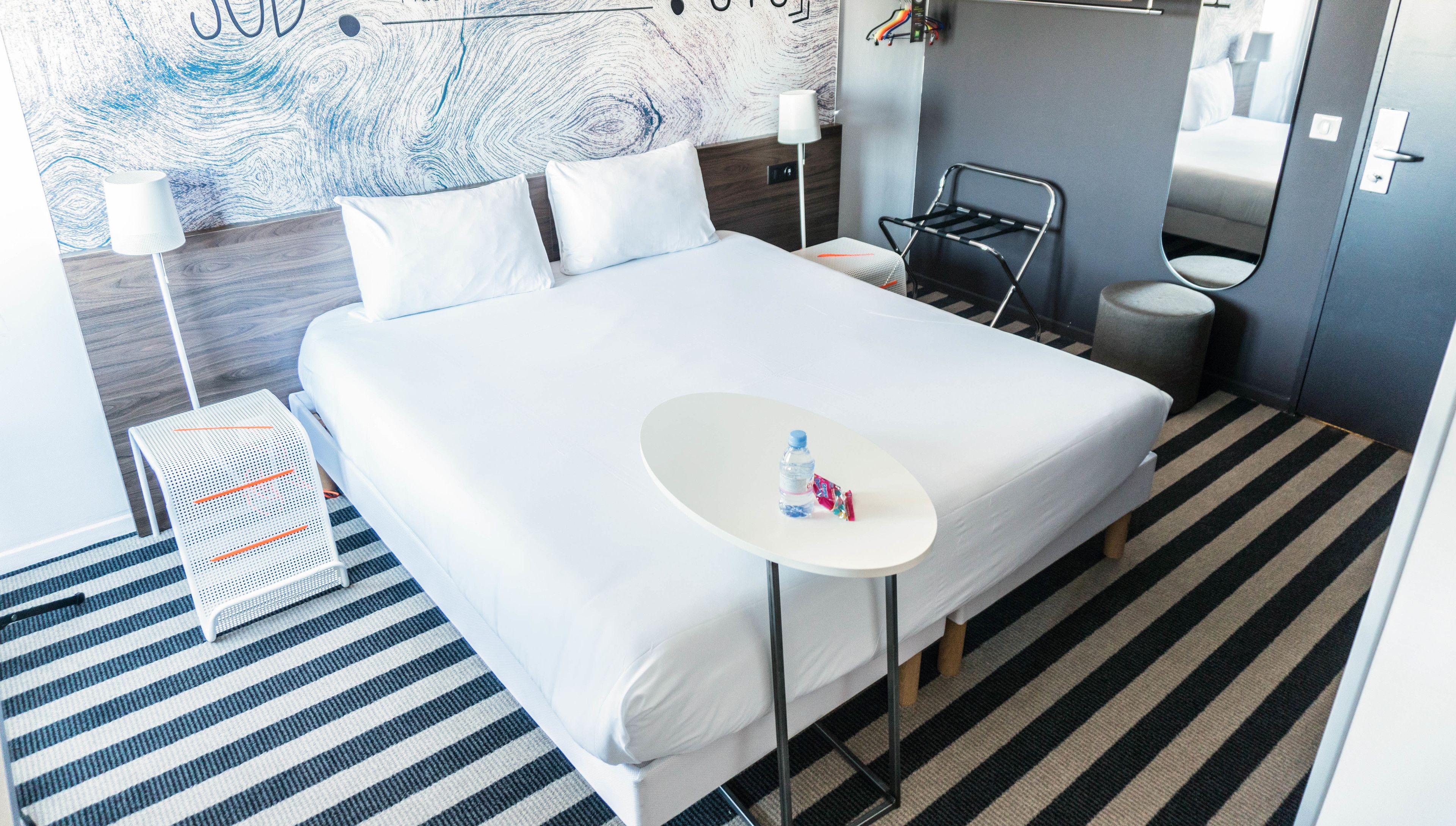 Ein Hotelzimmer mit einem großen Bett, einem kleinen runden Tisch mit einer Flasche und Snacks, einem Sessel, einer Lampe und einer Wand mit Holzoptik.