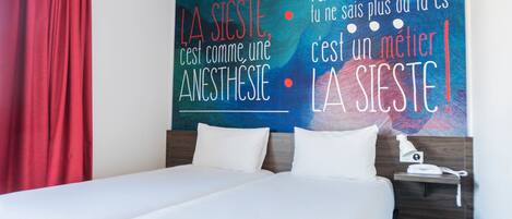 A hotel room with a bed, white bedding, a nightstand with a lamp, and a wall with motivational text in French.