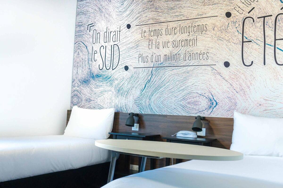 A hotel room with two beds, a small table, and a wall with French text.